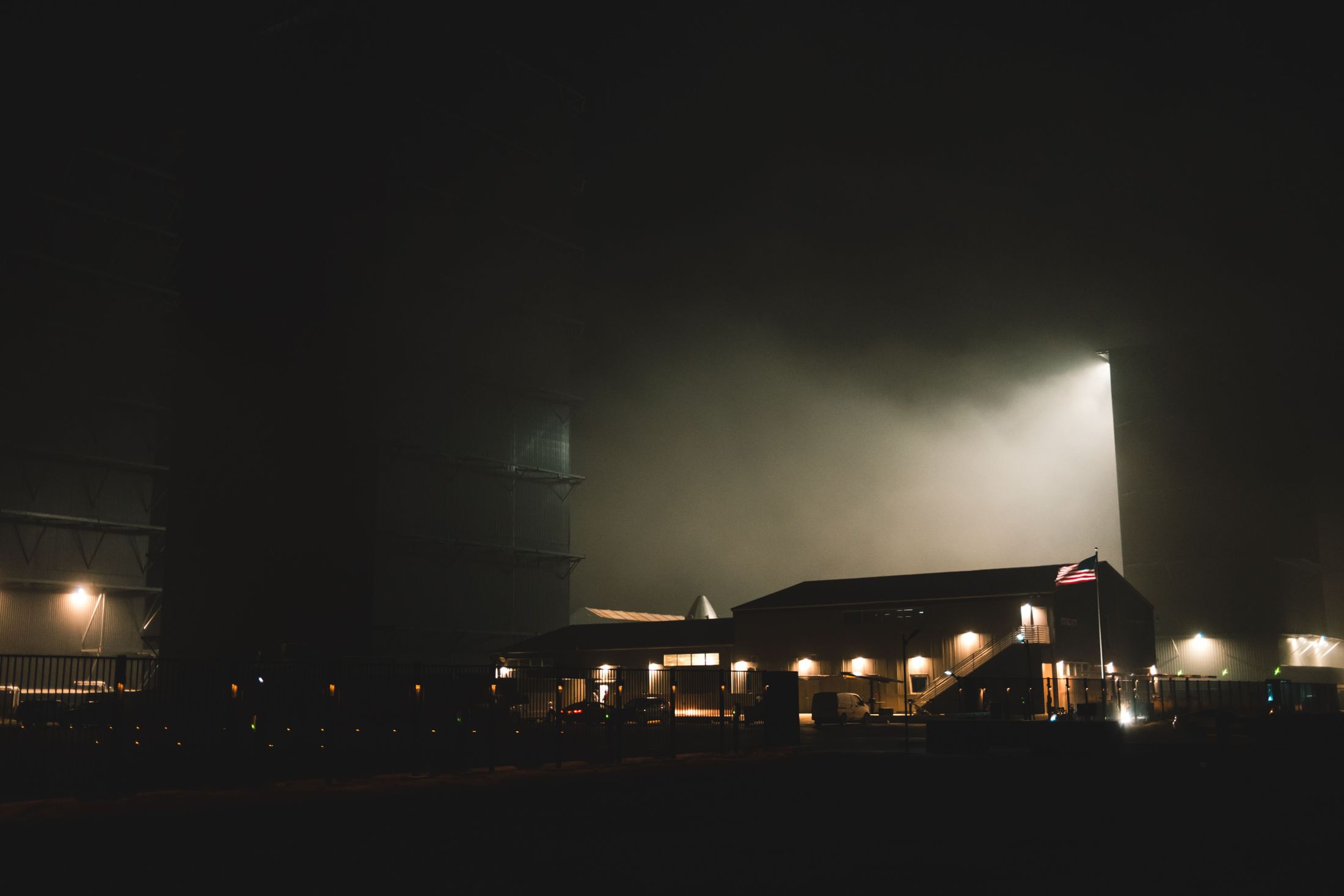 An industrial building at night, floodlit, its heat and fog rising into the sky, an American flag on a pole