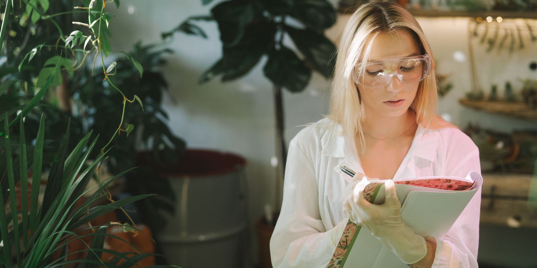 A technician in safety glasses takes notes among plants in a greenhouse